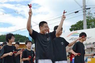 黒いTシャツを着た中学生くらいの男の子たちが笑顔で鳴子を掲げながら、揃って踊っている写真