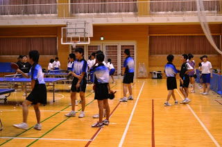 複数の中学生が体育館内でラケットを持って立っており、他の生徒の試合を見たり順番を待ったりしている練習風景の写真