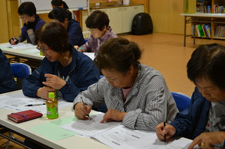 机に向かって座った女性たちが配布資料を見ながらボールペンで書き込み、真剣に学んでいる様子の写真