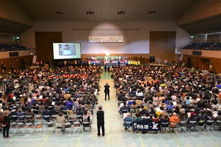 左側に大きなモニターがあり中央の花道に2名の人が立ち、両側にたくさんの人達が椅子に座り舞台に照明があたっている会場の全体を後方から写した写真