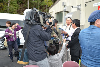 テレビカメラやマイクを持った報道関係者が、ふ化場の前で作業服やスーツ姿の関係者にインタビューしている様子を写した写真