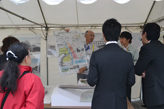 壁に建築図面などが掲示され、手前のテーブルには建造物の模型が置かれているテントの下で、大きな図面を広げて持つ男性の話を、周りに集まった複数の人々が聞いている様子の写真