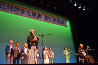 ステージで男性が尺八を吹き、もう一人が三味線を弾いている演奏に合わせて、後方に並んでいる年配の方々が歌を歌っている様子の写真