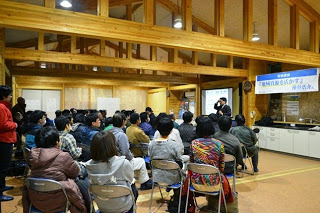 木造の建物内で多くの人々が椅子に座り、前方のスクリーンと講師に注目している様子が写され、「地域自然観察会のすすめ 丹沢大山」と書かれた横断幕が掲げられている写真