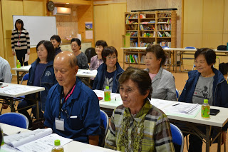 机に並んで座った参加者たちが前方を見ながら真剣な表情で話を聞いている様子の写真