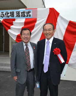 赤と白の垂れ幕の前で、スーツを着た男性二人が笑顔で並び、右の男性は胸に赤いリボンを付けて式典用の資料を持っている、施設完成記念式典での記念撮影の写真