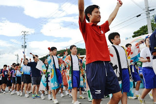 赤いシャツの少年を先頭に、カラフルな法被やスポーツウエアを着た多くの学生が隊列を組んでにぎやかに踊っている写真