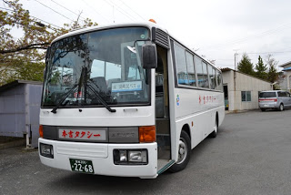 白い中型バスが建物の前に停まっており、側面に「登米タクシー」と書かれていて、正面からバスの全体像が見えるバス外観の写真