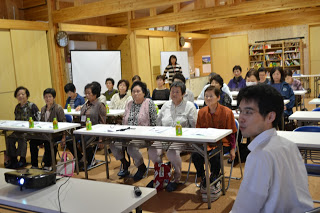 木造の室内で多くの参加者が正面のスクリーンを見つめ、講師が前方で話している場面を後方から撮影した写真