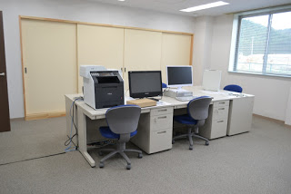 窓から光が差し込む室内で、白いデスクと青い椅子、パソコンやプリンターなどが整然と並ぶ南三陸町小森ふ化場の事務室の写真