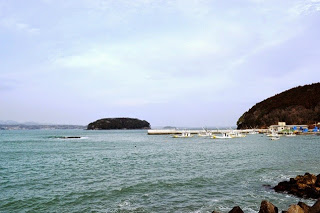 穏やかな海の先に小さな島が浮かび、防波堤や停泊中の漁船、右側には山も見える写真