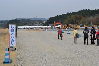 水色と白の配色のカラーコーンが設置され、左端に「しおさい通り」と書かれた看板が立っている砂利の広場を複数の人々が見学しており、奥にはショベルカーやトラックなどが駐車してある広場と白いテントが見え、その先に緑の山々が見える写真