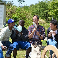 屋外のベンチに外国人を含む4人男性が座っており、それぞれ綿あめを楽しそうに食べている写真