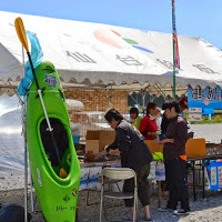 白いテントの下に複数の人々が集まっており、テント横には黄色いパドルが添えられた緑色のカヤックが立てかけられている写真