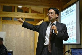 スーツ姿の藻谷浩介さんがマイクを持ってプレゼンテーションをしており、背景にスライドが映し出されたスクリーンが見える様子を写した写真