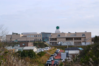 ベージュ色の大きな3階建ての施設の屋上に、緑色の十字のマークがある南三陸病院・総合ケアセンター南三陸の建物外観の写真