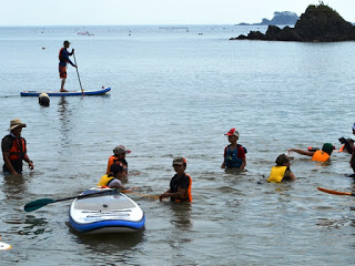 子どもたちが海の浅瀬に入って遊び、少し先にはパドルボートの上に立って水面を進んでいる人が見える写真