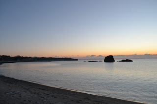 澄んだ空がオレンジから青へと移り変わる中、穏やかな海がその色を映し出し、岩が点在する静かな浜辺に波が優しく寄せる初日の出時の海岸の写真