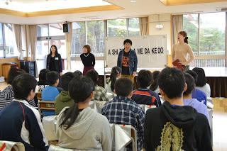 劇団四季の方々数名が教室の前に立って話しており、多くの児童が椅子に座って熱心に聞いている写真