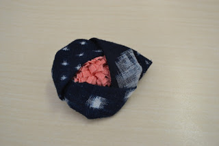 白い模様が入った濃い青色のカスリの内側にサーモンピンクの飾りが入った布細工のブローチの写真