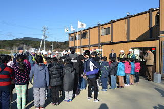 茶色い外壁の建設会社の建物の前で大勢の子どもたちと、ヘルメットと安全ベストを着用している建設会社の職員が向かい合って立っている写真