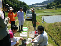 青空の下、田んぼのそばで生き物調査をしている人たちが机を囲んで男性の話を聞いている写真