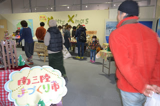 「南三陸 手づくり市」と書かれた看板が手前置かれた屋内の会場が多くの来場者で賑わっている様子の写真