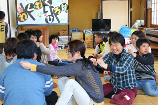 輪になって座った大人と子どもが背中をさすり合いながら、笑顔でふれあい遊びを楽しみ、和気あいあいと交流している様子を写した写真