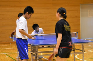 体育館の中で、4人の中学生が2対2のペアに分かれて卓球のダブルスの練習試合をしており、手前の男女2人がラケットを持って構え、奥の2人もそれぞれの位置についてプレーに集中していて、真剣な雰囲気の中で試合が進んでいる様子の写真