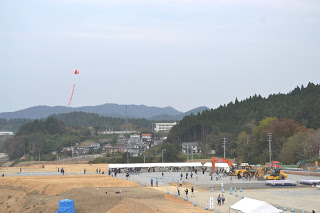 複数の重機が駐車されている平らに均された広い敷地に、白い屋根のテントが横一列に並び、たくさんの人々が分散して現場の見学を行っており、奥には緑の山々と斜面に建つ住宅等の建物が見え、空には宣伝の赤いバルーンが浮かんでいる志津川地区観光交流拠点整備予定地を遠くから撮影した写真