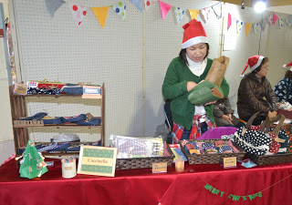 赤いテーブルクロスが敷かれたテーブルの上に布製品や小さなバッグなどの手作り品が並べられた販売ブースで、サンタ帽をかぶった女性が商品の陳列をしている様子の写真