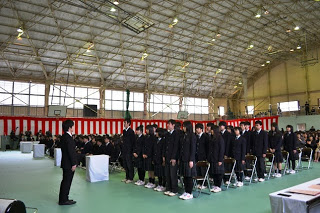 紅白幕が側面に飾られた天井が高く明るい体育館で、手前の席に座っていた制服姿の卒業生が、前方の先生と向かい合って立っている式典の様子の写真