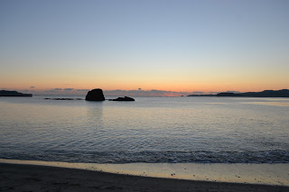 青からオレンジへと移り変わる空の下、穏やかな波が静かな浜辺に寄せる中、遠くに小さな島や岩が水面から顔を出す初日の出時の静かな海岸の写真