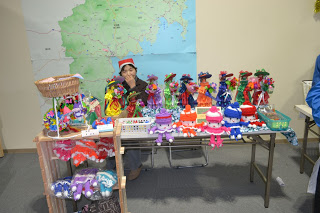 大きな地図が貼られた壁の前に、テーブルの上に色とりどりの手作りの人形が整然と並べられており、テーブルの左前には小さいクラフト作品が入ったバスケットが置かれた木の棚がある販売ブースの写真