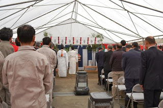 白い天幕の大きなテントの中で、ストーブや椅子が並べられた会場に人々が集まり、祭壇の前で白装束の人物が儀式を執り行っている安全祈願祭の様子の写真