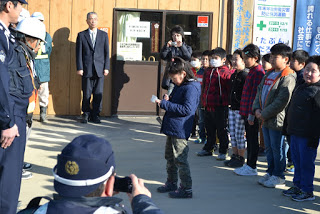 建物の前で複数の警察官に向かって一列に並んだ子どもたちのうち、1人がマイクを持って話し、周囲にはスーツ姿の大人や写真を撮る人がいる様子の写真