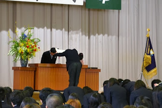 白幕や旗が飾られたステージ上で、壇上の演台に立つ先生に向かって、深くお辞儀をする生徒代表の姿が写された厳かな式典の写真
