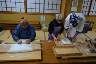 畳の床と障子がある和風の部屋で、2人の参加者が木のテーブルの上で麺棒を使って生地を伸ばしている様子が写されている、蕎麦打ち体験の写真