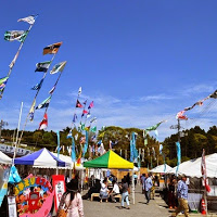 たくさんのテントや屋台が並び、青空にはたくさんのカラフルな旗がはためく屋外のイベント会場が、たくさんの来場者で賑わっている様子の写真