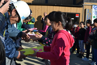 屋外で子どもたちが色とりどりの紙をヘルメットと安全ベストを着た建設作業員と思われる大人たちに手渡している様子の写真