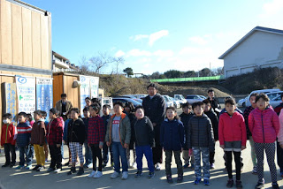 青空が広がり、背景には車が並んでいる建設現場の事務所の前で、冬服を着た子どもたちが整列している様子の写真