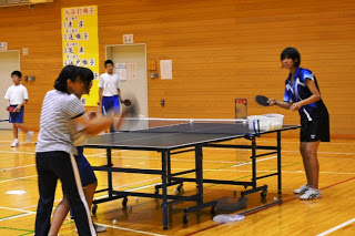 体育館の卓球台の前で、左側の生徒が指導者からラケットの正しい振り方を体に手を添えられて教わっている様子の写真