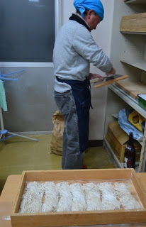 手前には麺の束のようなものが並んだ木製のトレーがあり、青い頭布、グレーのセーター、黒いエプロンを着た男性が、右奥の棚にそば切りの道具を直している様子の写真