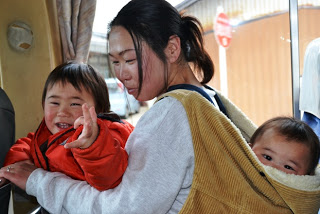 赤いジャンパーを着た幼い男の子が笑顔でカメラを見つめ、母親に背負われた赤ちゃんもこちらを見ており、親子3人がバス車内で移動している様子の家族の写真