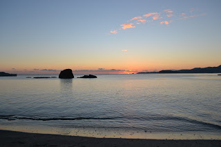 青からオレンジへと染まる空の下、雲が点在し、穏やかな海が朝焼けの色を映し出し、遠くには岩や陸地のシルエットが見え、手前では波が静かに砂浜に打ち寄せる朝方の海岸の写真
