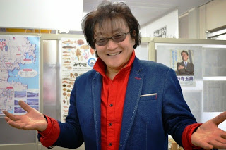 サングラスをかけ、青いジャケットと赤いシャツを着た亜KIRAさんが両手を広げて笑顔で写っている写真