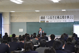 教室の前方で「講義『ふるさと南三陸を学ぶ』」と書かれた横断幕の下、黒板の前に立つ佐藤町長の話を、多くの学生が座って聞いている講話の様子の写真