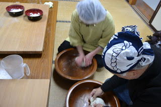 畳のある和風の室内で、テーブルには白い粉の入った赤いボウルが2つと透明のピッチャーが置かれている傍で、頭に三角巾をかぶった2人が床に座り、大きな木のボウルで生地をこねている写真