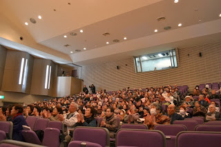 客席いっぱいに観客が座って舞台の方を向きながら拍手しており、天井が高く音響設備の整った明るいホールで行われている、発表会の観客席の写真