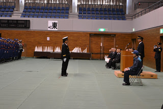 ベイサイドアリーナの屋内会場で、制服を着た隊員が中央に立って敬礼し、壇上の関係者や来賓が見守る中で人員報告が行われている出初式の写真